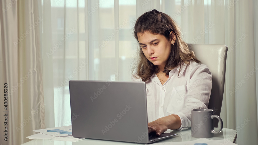 Disabled woman freelancer having cerebral palsy takes cup with problem to drink tea working from home on computer sitting at table against window. Disabled woman freelancer having cerebral palsy takes cup with problem to drink tea working from home on computer sitting at table against window.