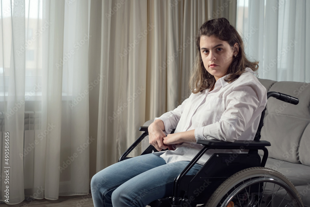Sad disabled woman with organic damage of central nervous system sits in wheelchair looking at camera behind curtain in living room at home. Sad disabled woman with organic damage of central nervous system sits in wheelchair looking at camera behind curtain in living room at home.