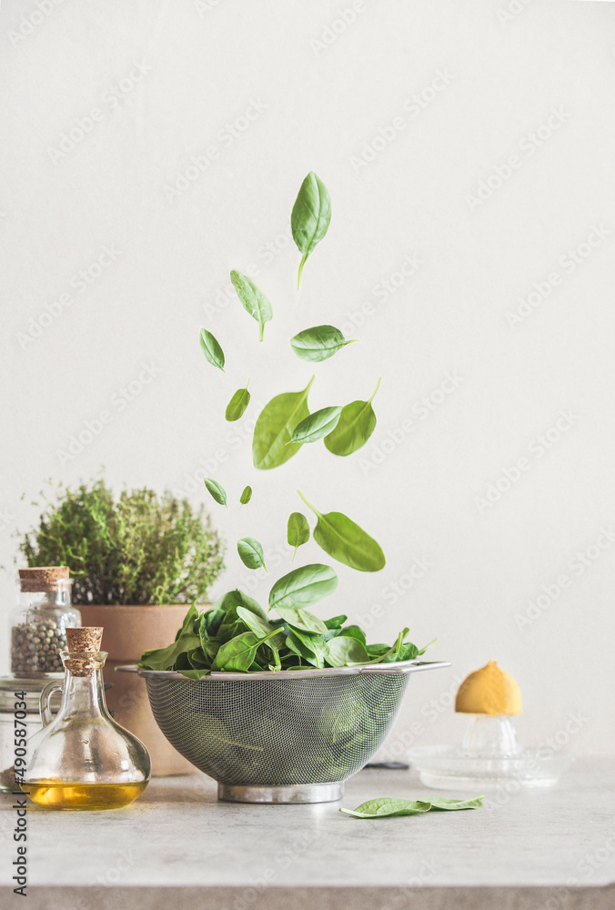 Falling green spinach leaves in sieve on kitchen table with ingredients:  olive oil, potted herbs, lemon and pepper. Food levitation concept. Healthy cooking preparation at home. Front view.