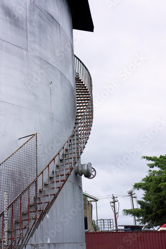 Rusty stairs and handrailings winding around a circular silo against a cloudy grey sky.