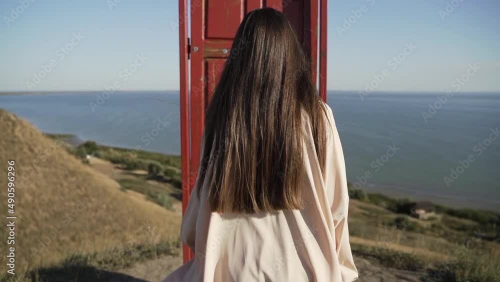 Young brunette with long hair in light clothes goes to an old red door on cliff in front of the sea in summer Young brunette with long hair in light clothes goes to an old red door on cliff in front of the sea in summer