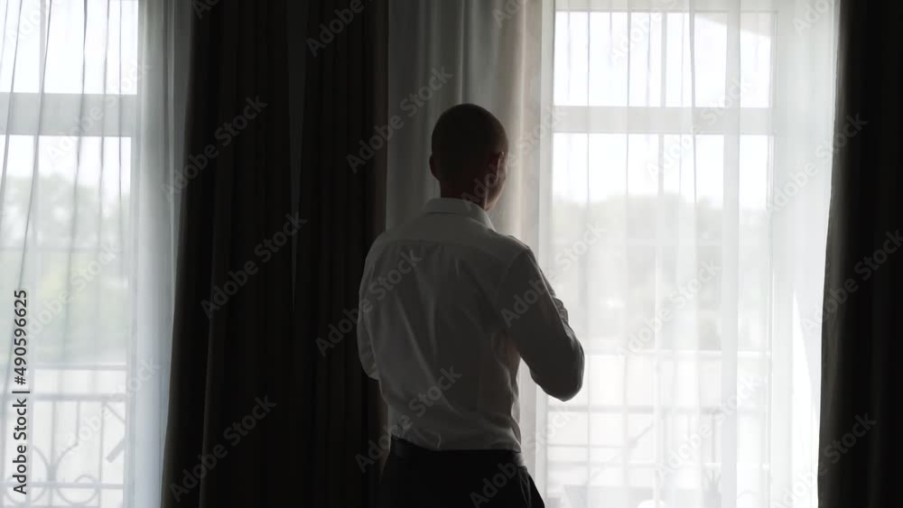 Man in black trousers and white shirt stands near the window and tightens button on his collar. Groom gets dressed for the wedding in the morning in the room Man in black trousers and white shirt stands near the window and tightens button on his collar. Groom gets dressed for the wedding in the morning in the room