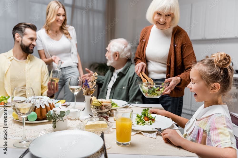 senior woman holding vegetable salad near granddaughter and blurred family.