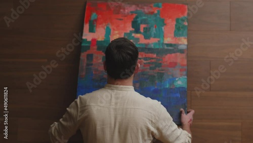 Contemporary art. Stylish guy artist hang colourful abstract artwork on wall, rear view, handheld video. ProRes codec