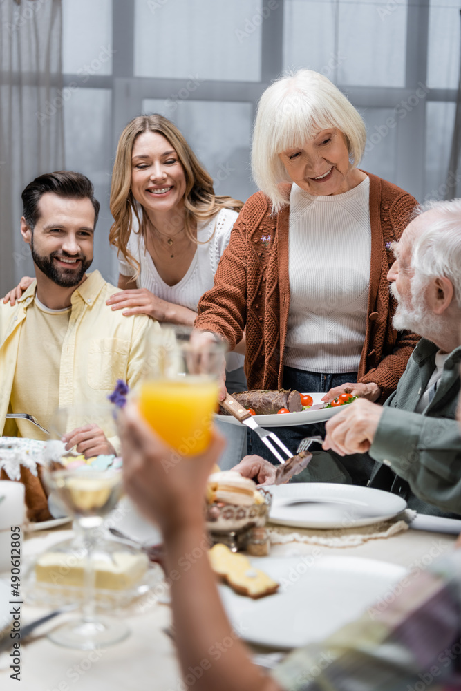 smiling senior woman serving fried meat near family and blurred kid with glass of orange juice. smiling senior woman serving fried meat near family and blurred kid with glass of orange juice.