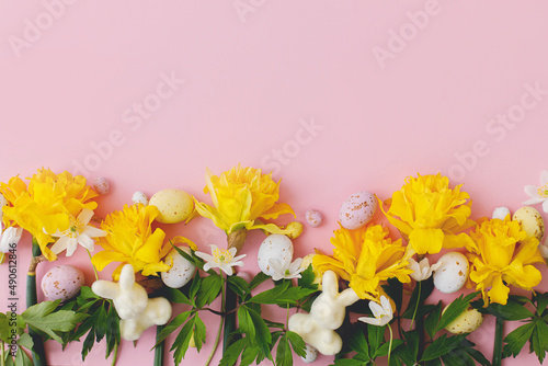 Colorful Easter chocolate eggs, bunnies and daffodils flowers border on pink background. Happy Easter! Stylish Easter flat lay. Greeting card or banner template