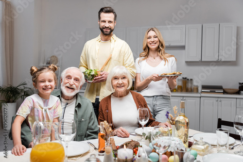 Canvas Print happy couple holding vegetable salad and cookies near family during easter celebration