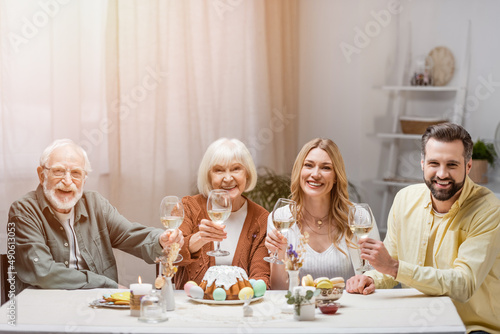 pleased family holding wine glasses and smiling at camera near easter cake and painted eggs.
