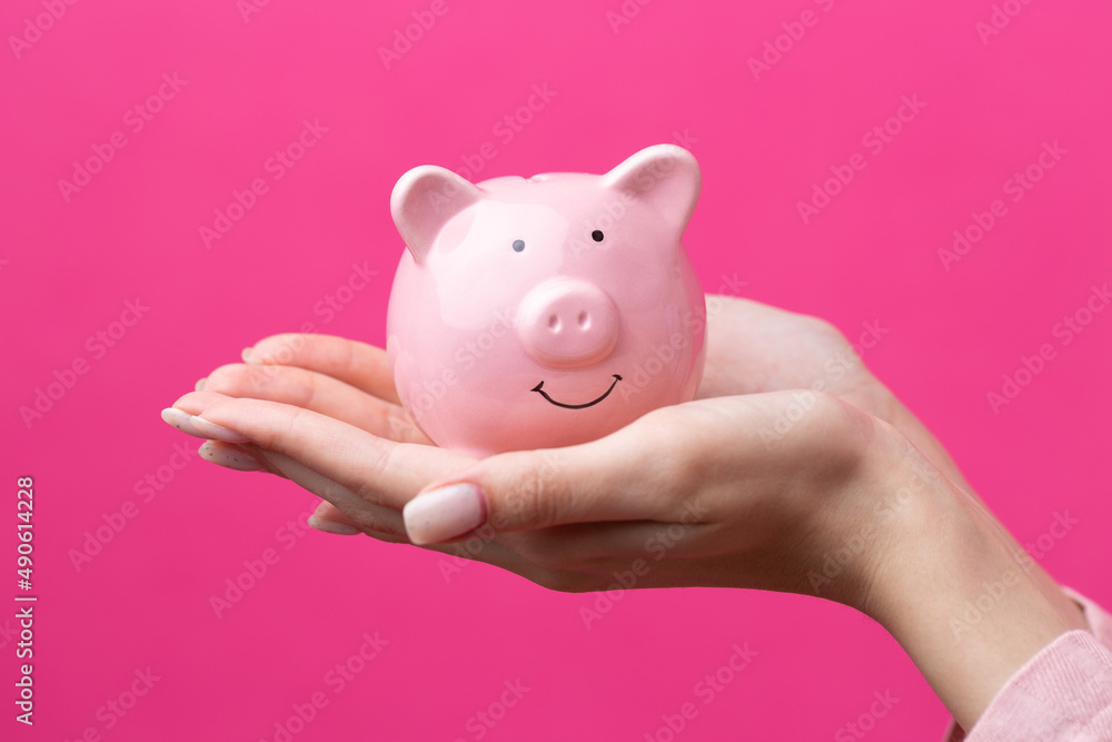 A cute girl in a pink jacket is holding a 3d piggy bank in her hands. Beautiful close-up portrait in studio on pink.