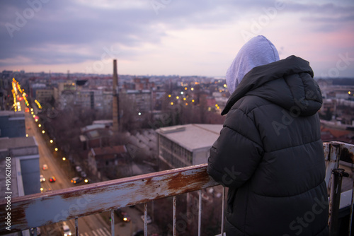 Depressed sad young female in jacket with hoodie standing on the edge of the fence on residential building rooftop. Suicide and major depressive disorder concept. Contemplating suicide.