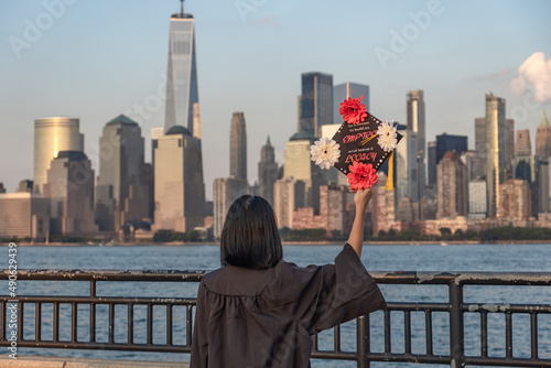 A girl stands on the New Jersey side of the Hudson River holding her graduation cap and wearing her graduation gown with downtown Manhattan in the background. 