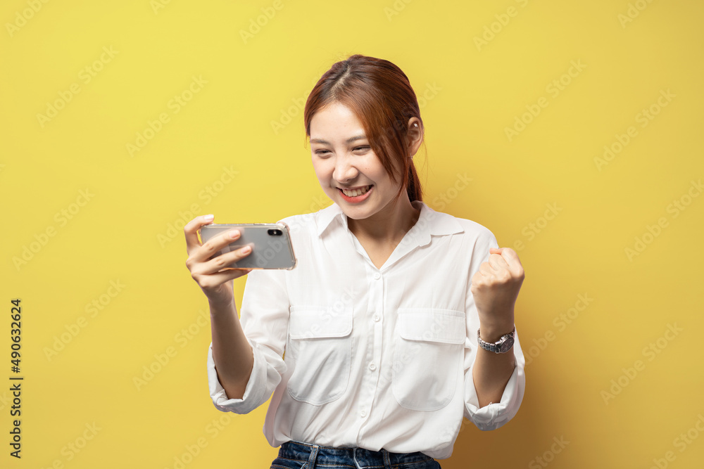 Success. Excited young woman playing mobile video game, holding smartphone horizontally and smiling amused, winning online, standing in shirt against yellow background. Success. Excited young woman playing mobile video game, holding smartphone horizontally and smiling amused, winning online, standing in shirt against yellow background.