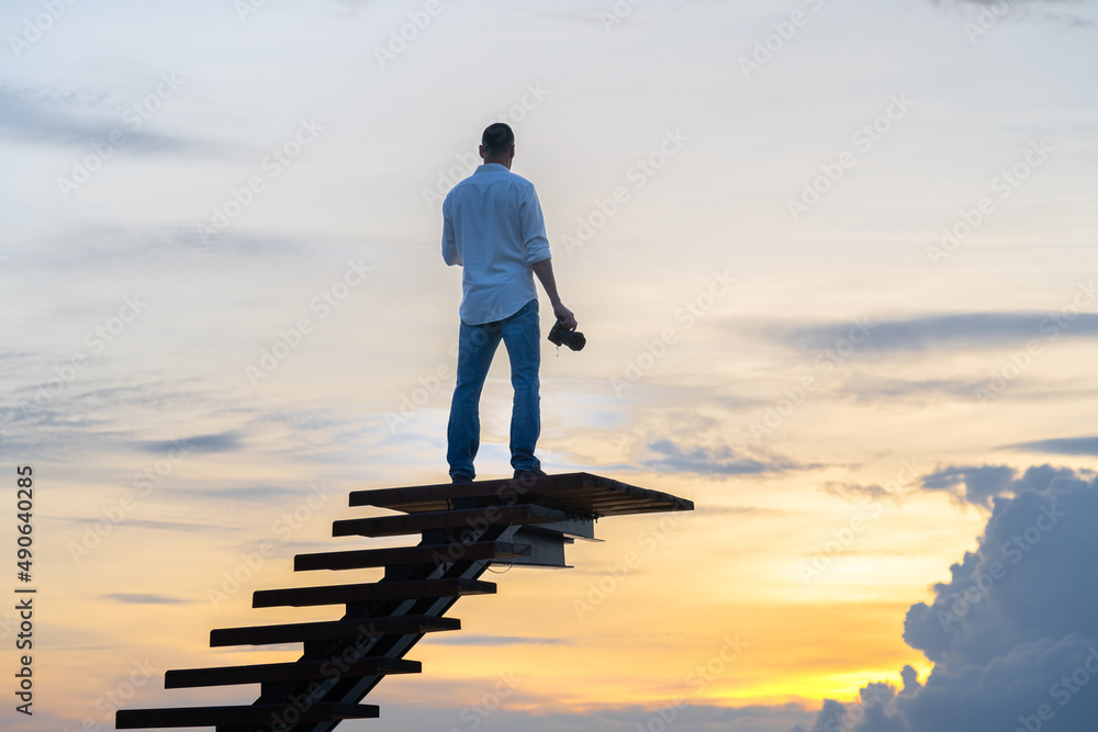 A man in jeans and a white shirt is standing on the stairs against the colorful sky with a camera in his hand