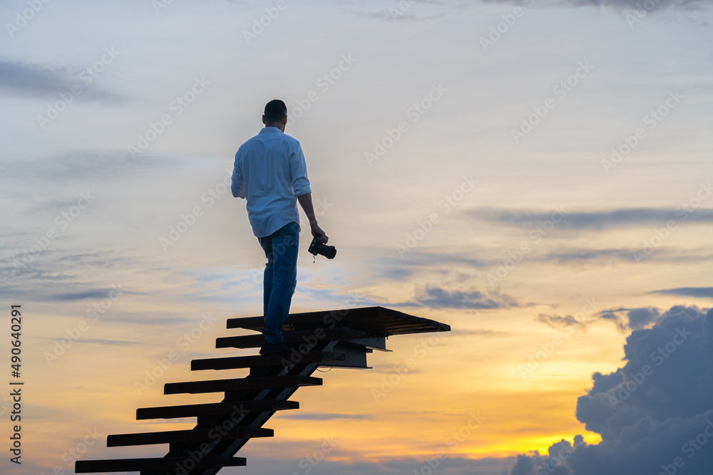 A man in jeans and a white shirt is standing on the stairs against the colorful sky with a camera in his hand