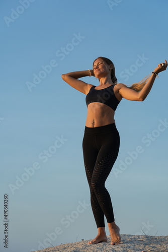 Fototapeta Naklejka Na Ścianę i Meble -  Young woman in sportswear with long hair standing on the rock by the sea and enjoying sunrise on a summer morning. Blue sky on the background.