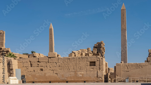 Hieroglyphs and carvings are visible on the dilapidated walls of the Karnak Temple in Luxor. Two obelisks rise against a clear blue sky. Egypt