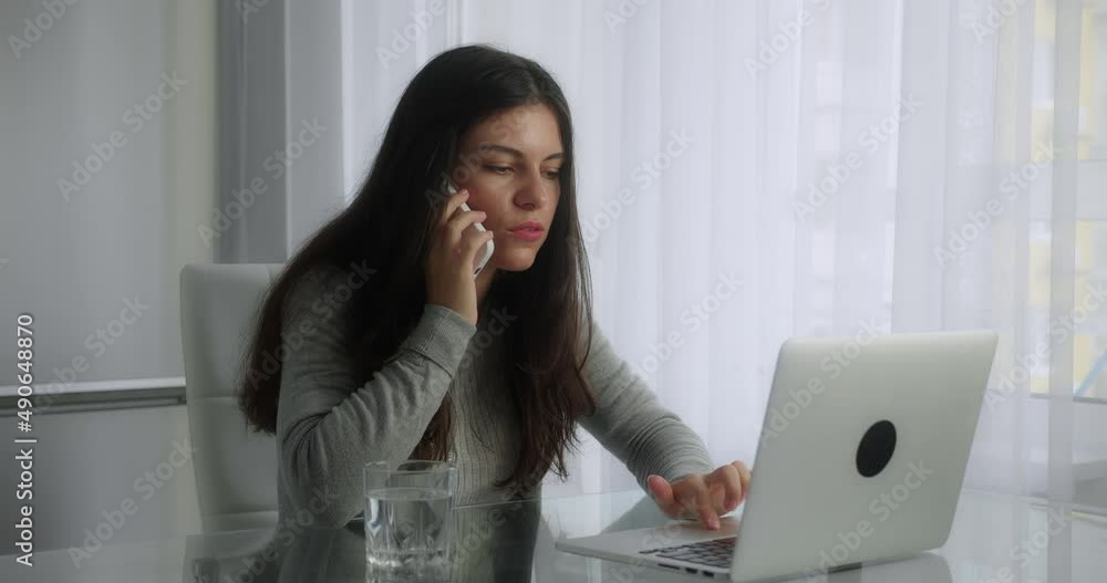 Young busy brunette business woman at her desk, talking on the phone and looking at computer of the modern home office background Young busy brunette business woman at her desk, talking on the phone and looking at computer of the modern home office background