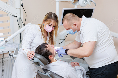 The dentist doctor looks at the patient s teeth and holds dental instruments near the mouth. The assistant helps the doctor. They wear white uniforms with masks and gloves. Dentist. Dental office