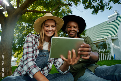 Mixed race male and female famers sitting together on green grass looking at digital tablet photo