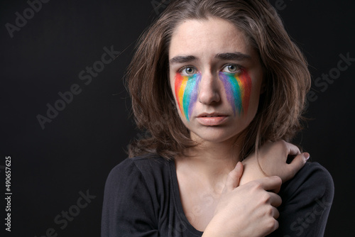 Dejected lesbian European appearance with colorful rainbow flag in form stripes on her cheek flowing from her eyes. Pride Month. LGBT girl on a black background in black clothes