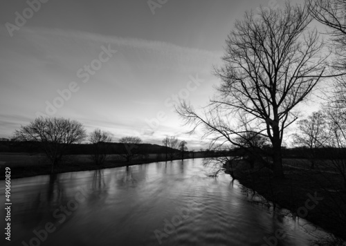 Evening winter sunset above River Ruhr near Schwerte Germany. Rural area with bare tree silhouettes reflected by the water surface. Black and white scenery panorama with high contrast.