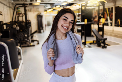 Fit young woman with a towel at health club. Female relaxing after workout. Portrait of charming smiling young fitness girl with a towel and posing while looking at the camera in the gym.