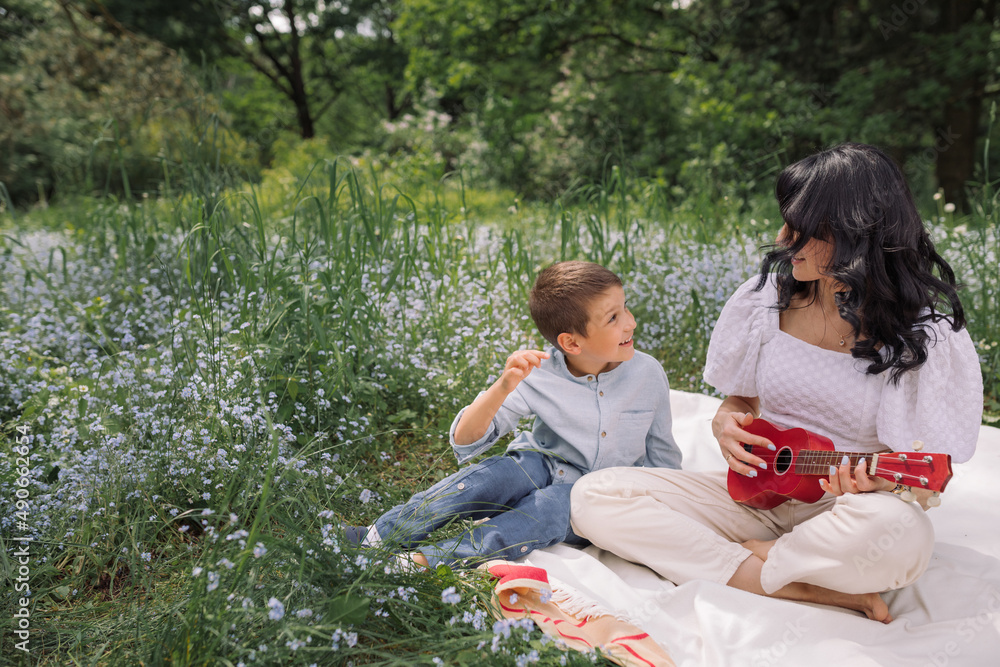 Mother singing playing on ukulele while sitting with her son on picnic blanket in the park. Mother singing playing on ukulele while sitting with her son on picnic blanket in the park.