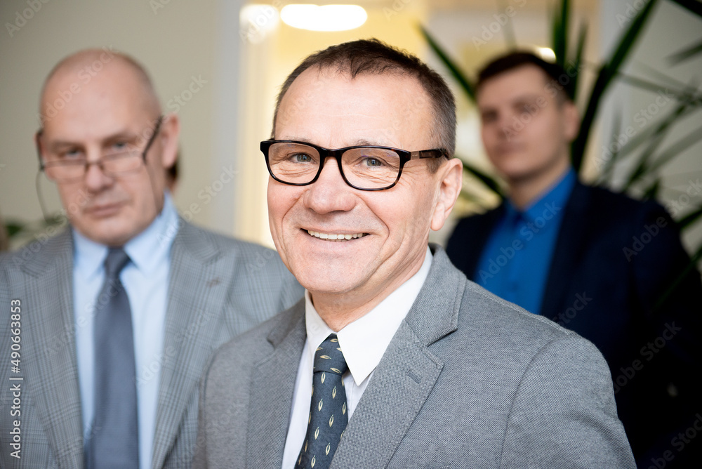 Portrait of smiling confident senior businessman standing with people of team in background Portrait of smiling confident senior businessman standing with people of team in background