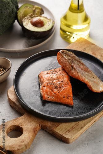 Two grilled fish steaks - red salmon on black round plate on wooden board, olive oil, avocado on grey background