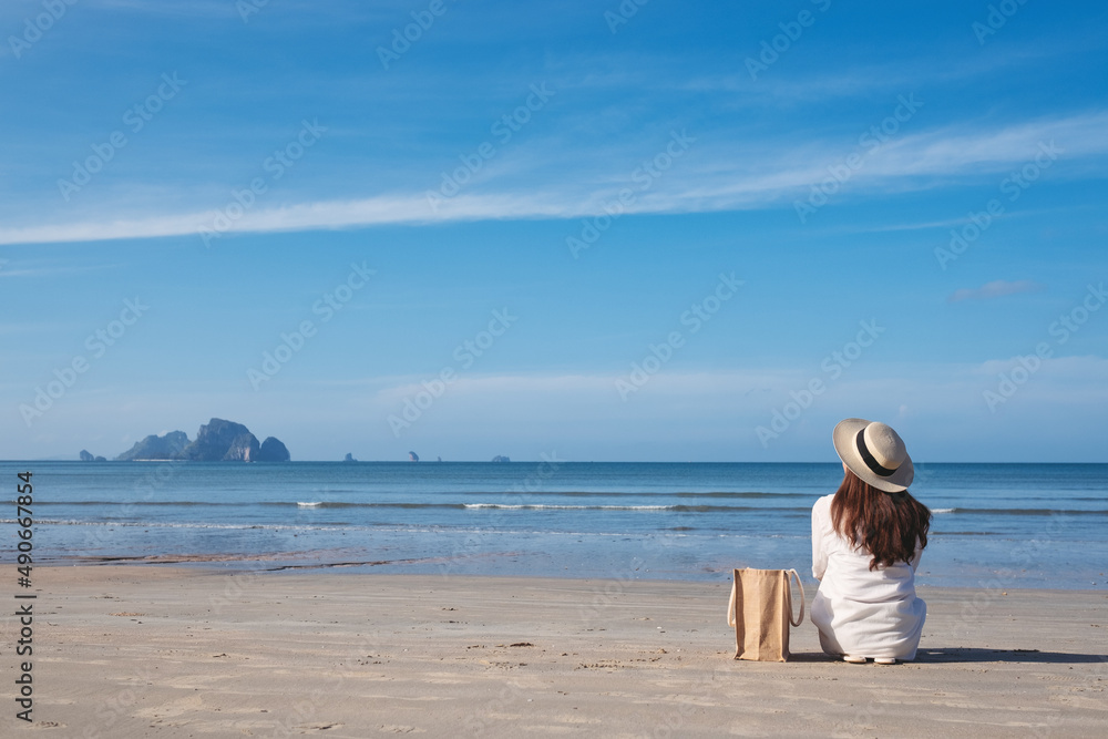 Rear view image of a young woman with hat and bag sitting on the beach with blue sky background