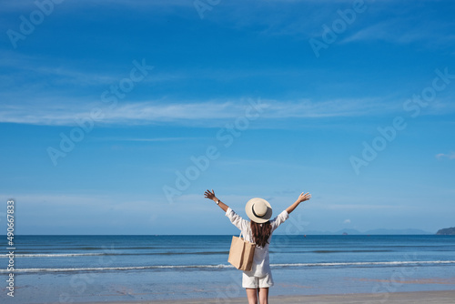 Rear view image of a woman with hat and bag opening arms while walking on the beach with blue sky background © Farknot Architect