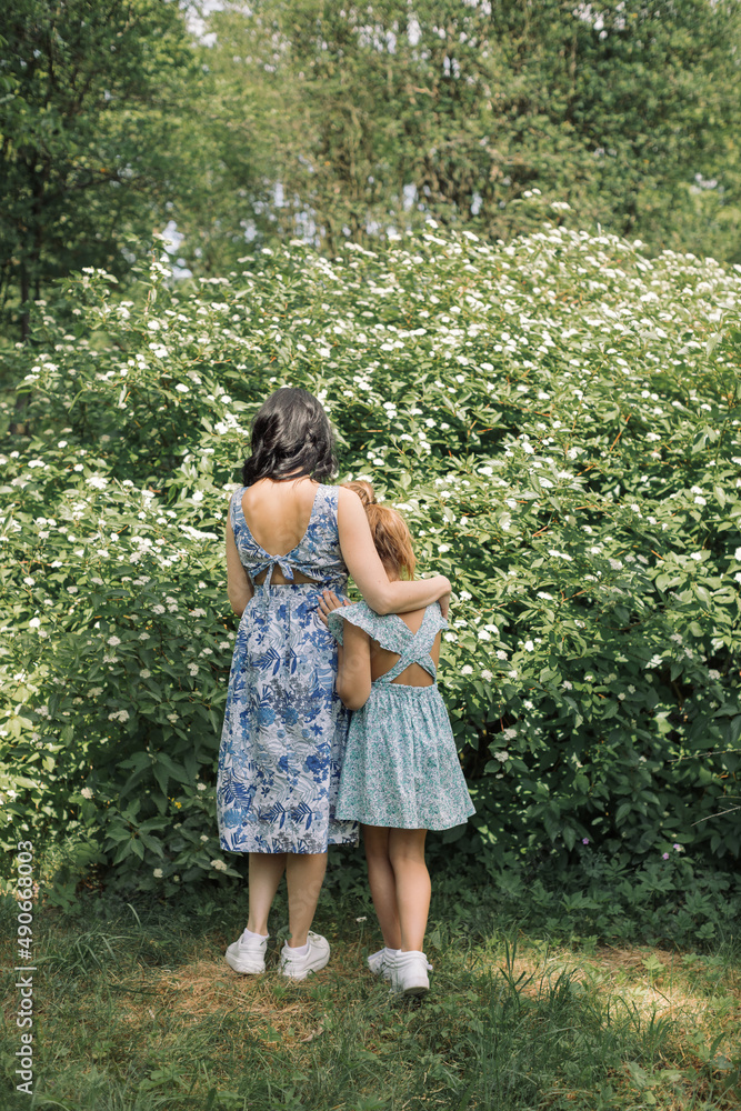 View from behind of a mother and daughter in floral summer dresses standing near bush in blossom. Childhood. View from behind of a mother and daughter in floral summer dresses standing near bush in blossom. Childhood.
