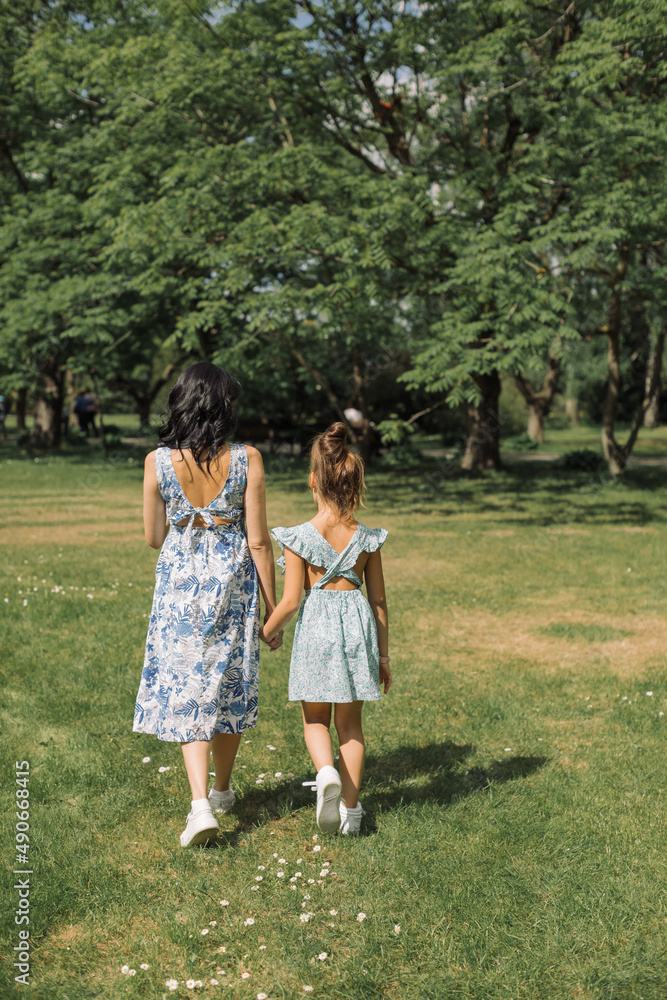 Young mother walking with her little girl in floral dress on a sunny day, having fun in the park.