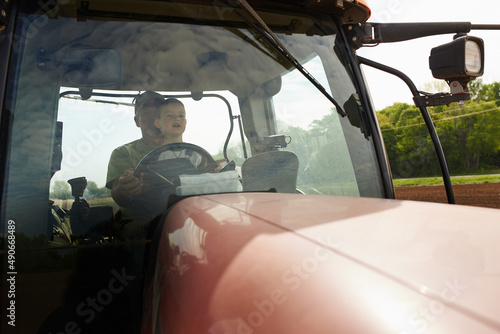 Work begins early on the farm. Shot of a young boy sitting with his dad inside the cab of a modern tractor.
