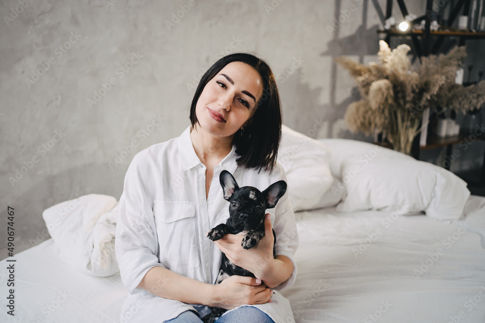 happy attractive brunette woman with a French Bulldog puppy in a bedroom.