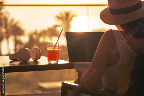 Silhouette of woman freelancer in straw hat drinking juice during working on laptop sitting on the armchair near table on balcony opposite sunset palm sea beach. Remote work on the vacation. Rear view photo