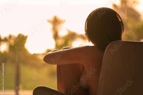 Rear view of naked woman in headphones hugging her knees sitting on chair near panoramic window and looking on palm trees sea beach at sunset. Female listenning music. photo