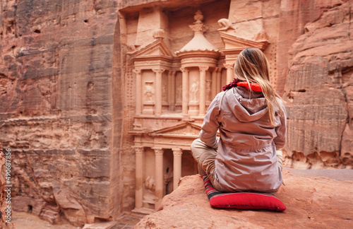 Young woman in winter jacket sits on small red pillow at viewpoint looking to Al-Khazneh - treausry, main attraction of Petra, Jordan. View from behind