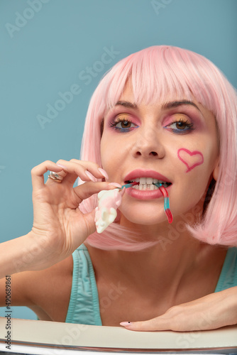 sexy glamour woman with fashionable makeup on pretty face and short hairstyle pink wig in dress eating marmalade mouse in studio on grey background  copy space