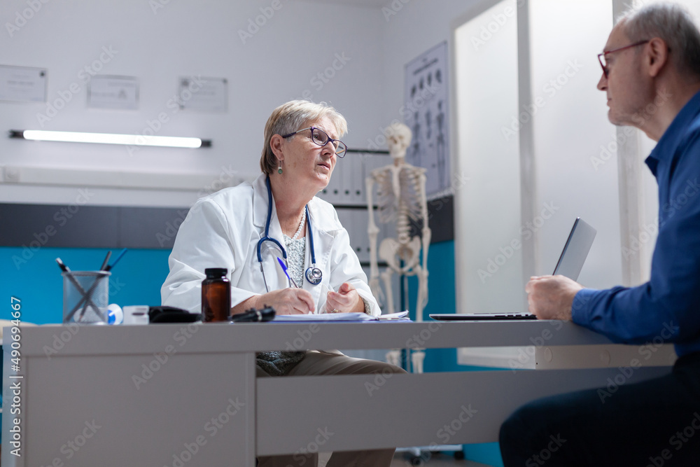 General practitioner consulting sick patient and preparing to sign checkup papers at medical appointment. Woman doctor having discussion with aged man about healthcare diagnosis.