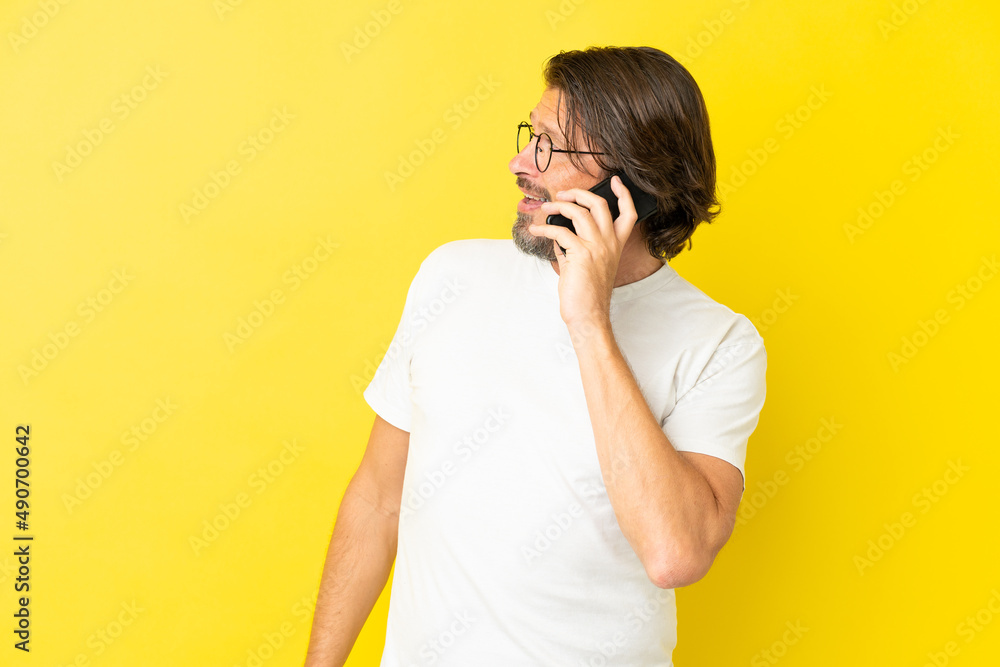Senior dutch man isolated on yellow background keeping a conversation with the mobile phone with someone Senior dutch man isolated on yellow background keeping a conversation with the mobile phone with someone