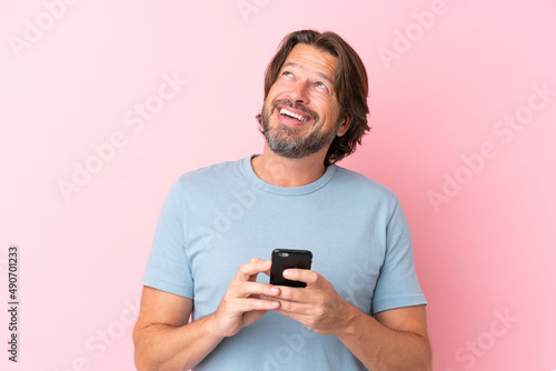 Senior dutch man isolated on pink background using mobile phone and looking up © luismolinero
