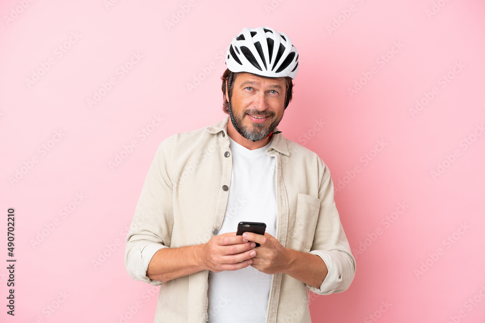 Senior dutch man with bike helmet isolated on pink background sending a message with the mobile Senior dutch man with bike helmet isolated on pink background sending a message with the mobile