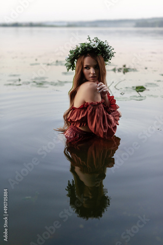 Mavka water nymph naiad near the water in nature forest on the river bank of the lake on a quarry on a cliff in a brown orange dress with a wreath on her head and candles in her hands Ivan Kupala photo