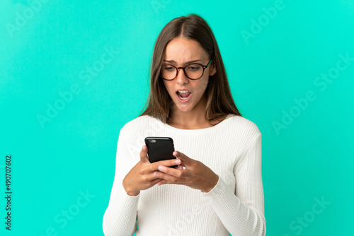 Young woman over isolated blue background looking at the camera while using the mobile with surprised expression