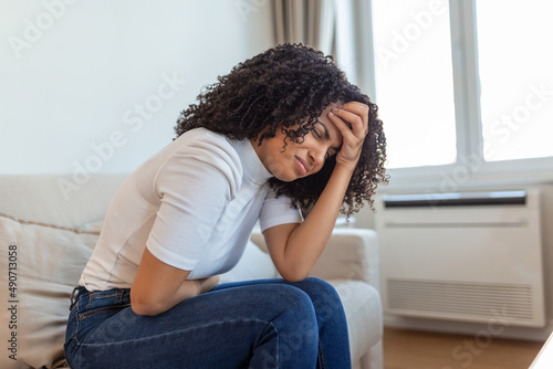 African American Woman in painful expression holding hands against belly suffering menstrual period pain, lying sad on home bed, having tummy cramp in female health concept