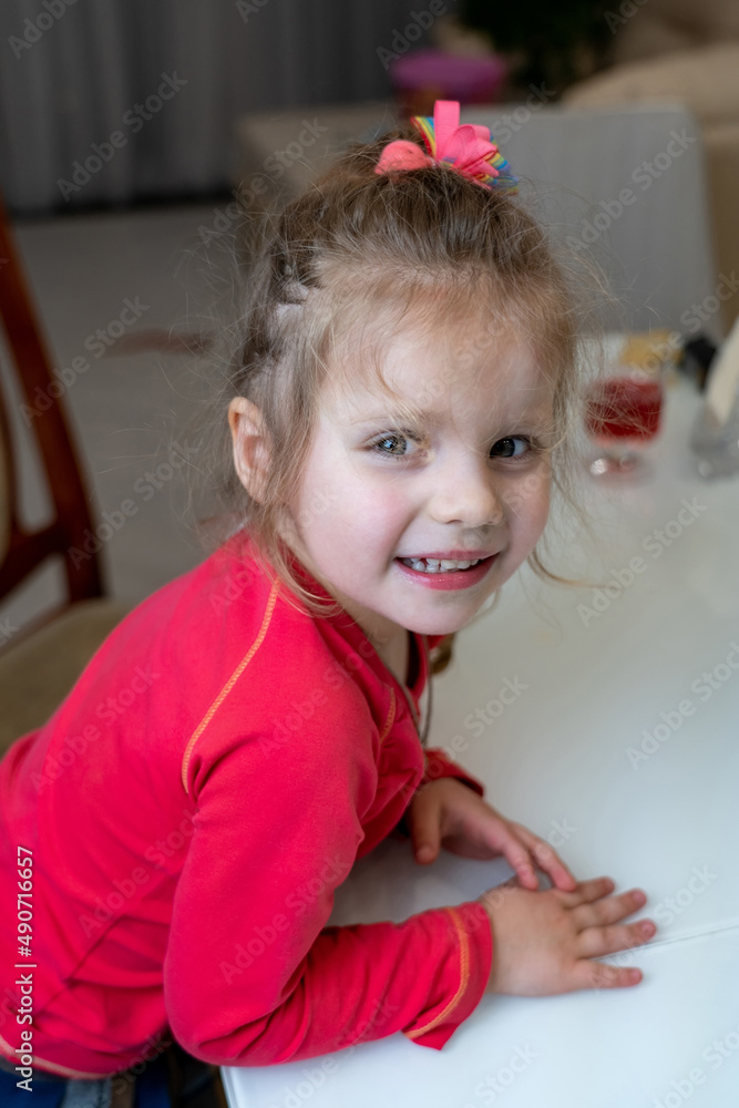 Charming little girl with blonde hair and colorful hairpins looks at the camera, smiles sweetly and feels good at table in the kitchen Charming little girl with blonde hair and colorful hairpins looks at the camera, smiles sweetly and feels good at table in the kitchen