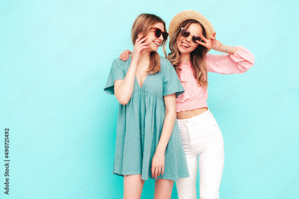 Two young beautiful smiling brunette hipster female in trendy summer dresses. Sexy carefree women posing near blue wall. Positive models having fun. Cheerful and happy. In hats and sunglasses Two young beautiful smiling brunette hipster female in trendy summer dresses. Sexy carefree women posing near blue wall. Positive models having fun. Cheerful and happy. In hats and sunglasses