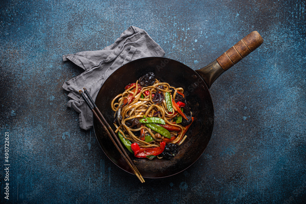 Asian dish stir fry udon noodles with vegetables and mushrooms in black rustic wok pan with wooden chopsticks on rustic dark blue concrete background from above, Chinese fast food - obrazy, fototapety, plakaty 