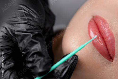 Master applies protect gel on red of lips young woman after permanent makeup tattoo in beautician salon